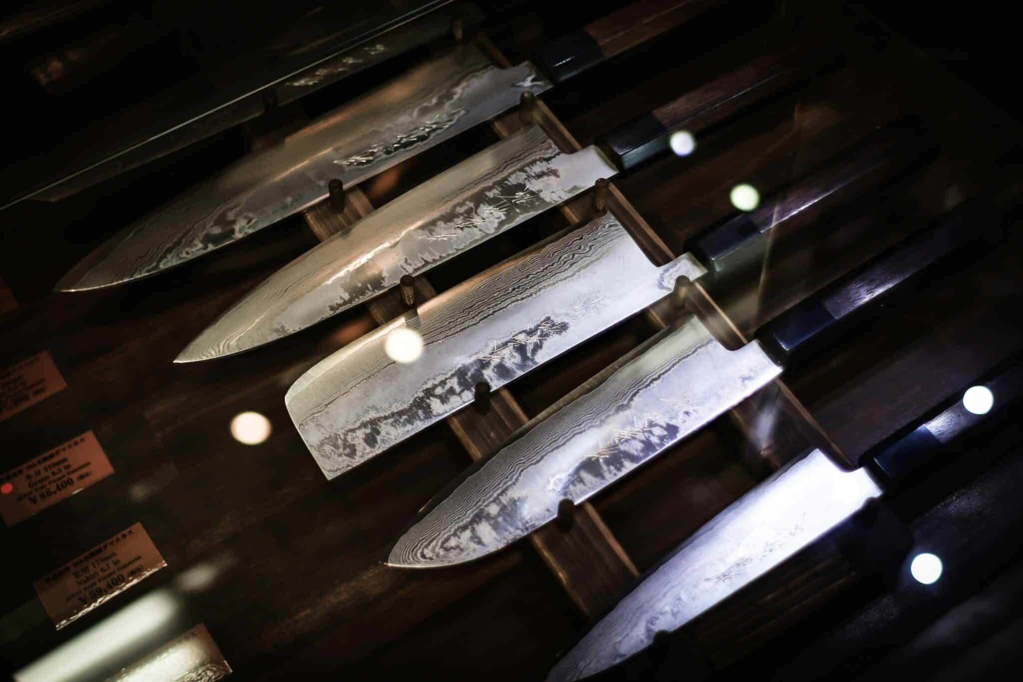 Et udvalg af forskellige typer japanske knive. Med traditionelt wa-håndtag. Liggende i en montre med prisskilte ved siden.